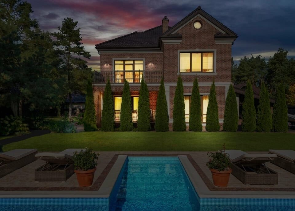 Brick two-story home at dusk with lit windows, a lawn and a long pool