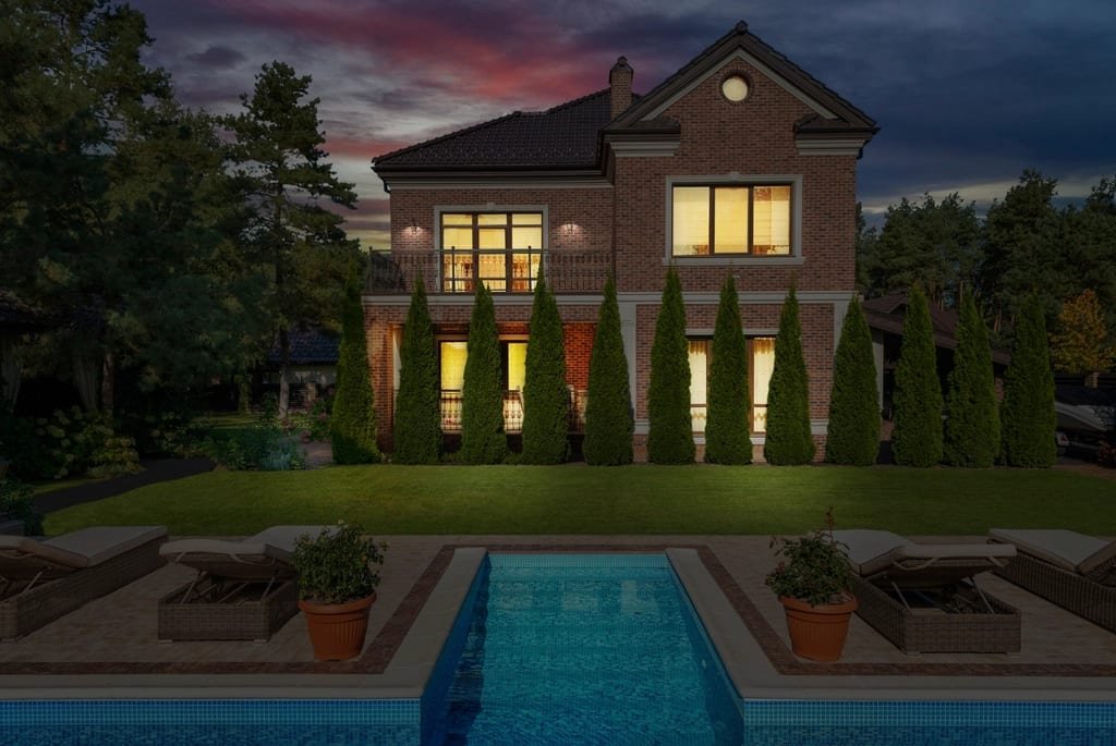 Brick two-story home at dusk with lit windows, a lawn and a long pool